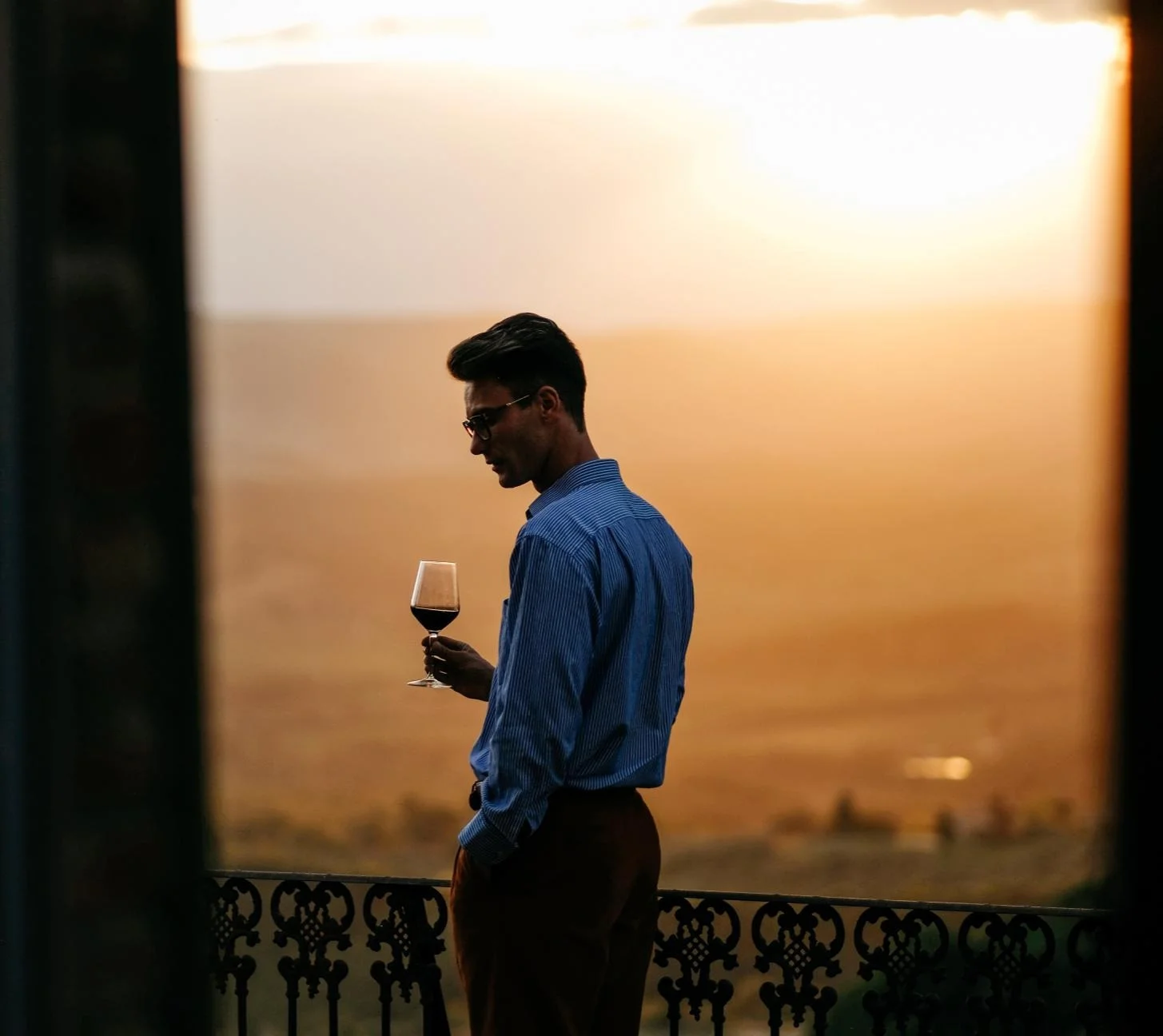 Un uomo di profilo osserva un calice di vino rosso affacciato su una terrazza al tramonto